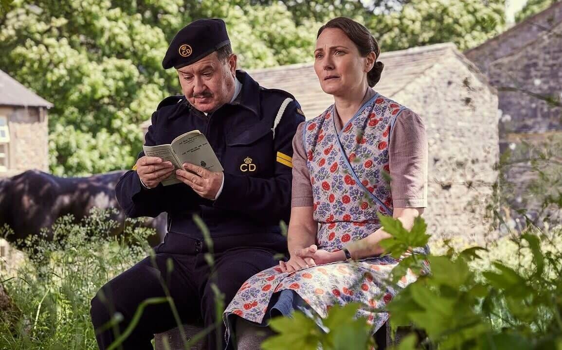 Mr. Bosworth (Jeremy Swift) in his uniform, reading an instruction manual, and Audrey Hall (Anna Madeley) sit outside together.