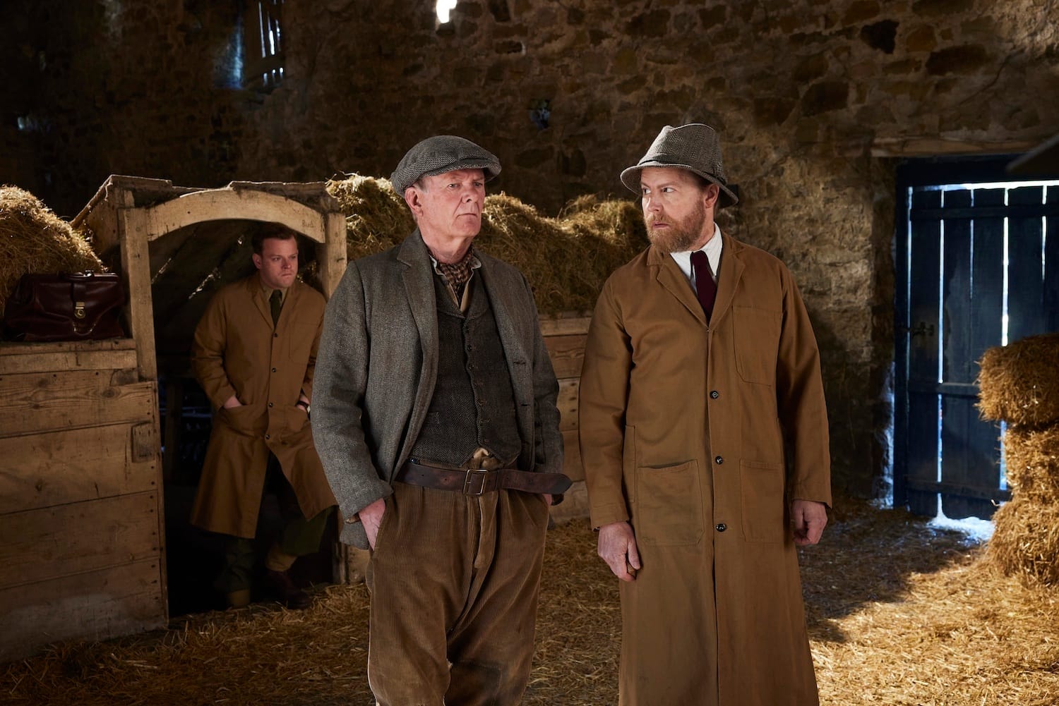 Tristan Farnon (Callum Woodhouse), Butch Fielding (Walt Bedford), and Siegfried Farnon (Samuel West) in the stable. The Farnon brothers look apprehensive about the procedure they're about to perform.