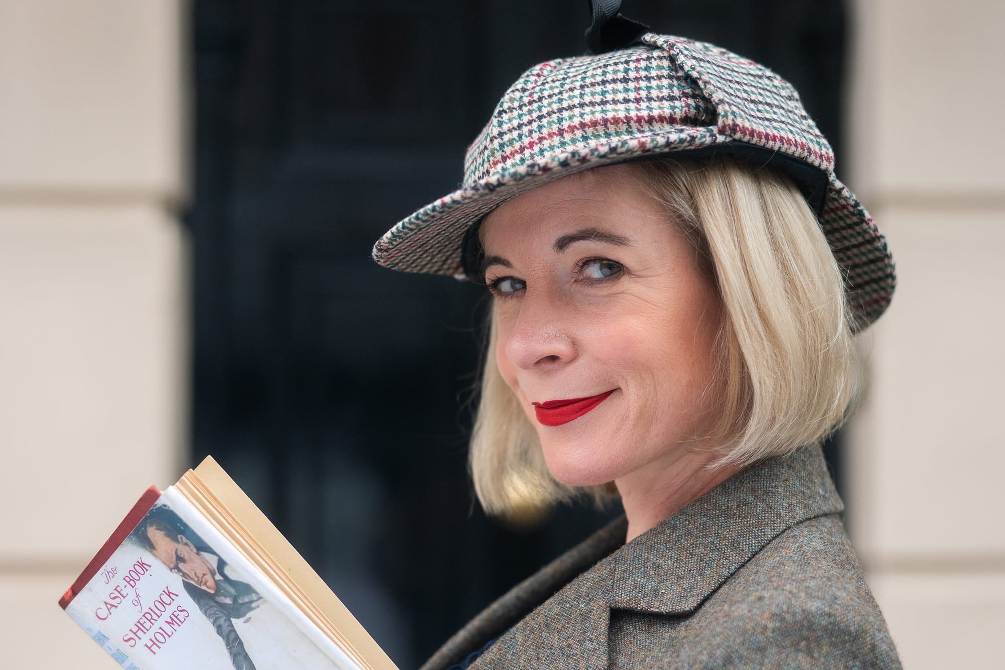 Lucy Worsley in a deerstalker hat and book in hand in front of the 221B Baker Street door in 'Lucy Worsley's Holmes vs. Doyle.'