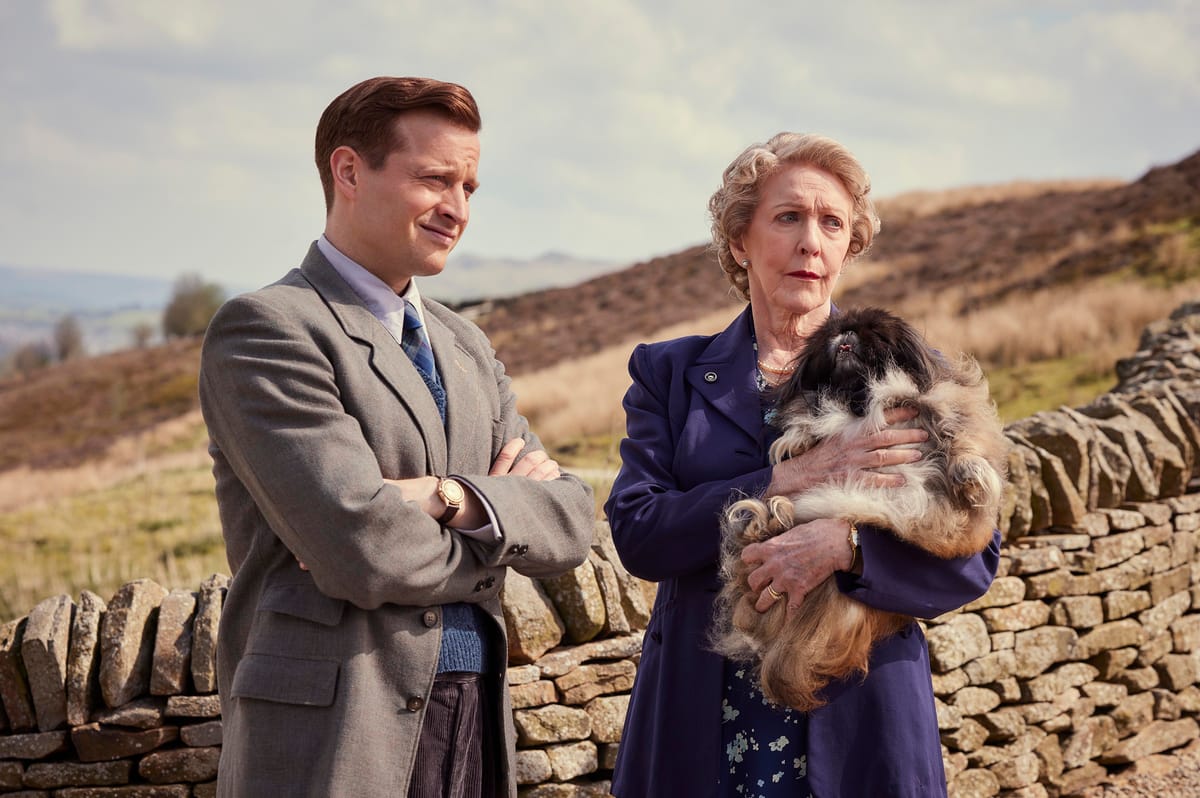 Standing in front of a drystone wall, James Herriot (Nicholas Ralph) with Mrs. Pumphrey (Patricia Hodge) and her beloved Pekinese Tricki Woo