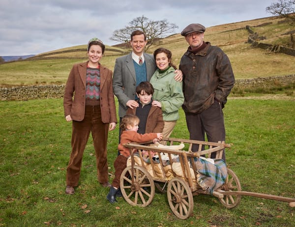 Standing in a green field and smiling, James and Helen Herriot, with their two small children Jimmy and Rosie, and Richard Alderson. The children have a miniature farm cart to ride in..